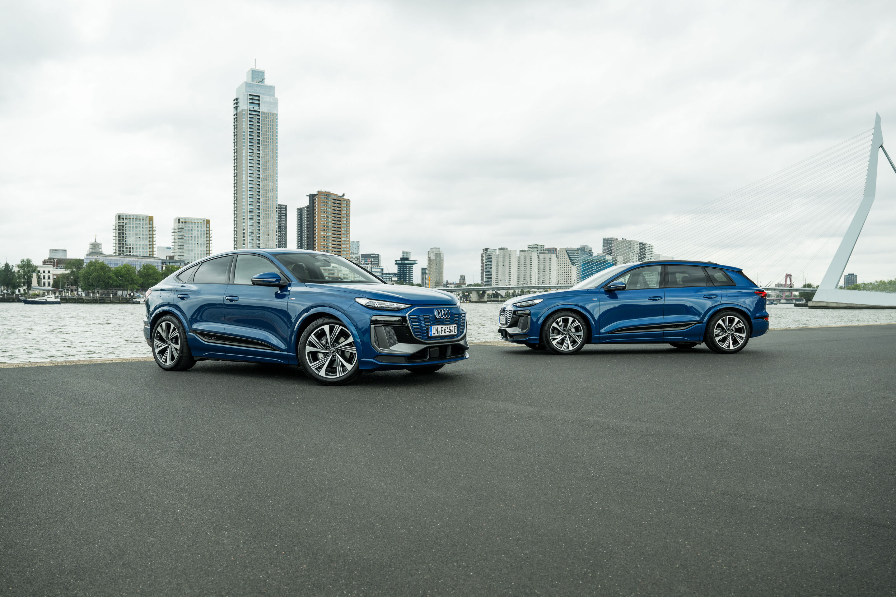 Audi Q6 e-tron Lifecycle Experience - Standaufnahme: Zwei Audi Q6 e-tron (Ascariblau), als SUV und Sportback, seitlich und schräg in urbaner Umgebung am Wasser mit moderner Skyline und Brücke im Hintergrund.