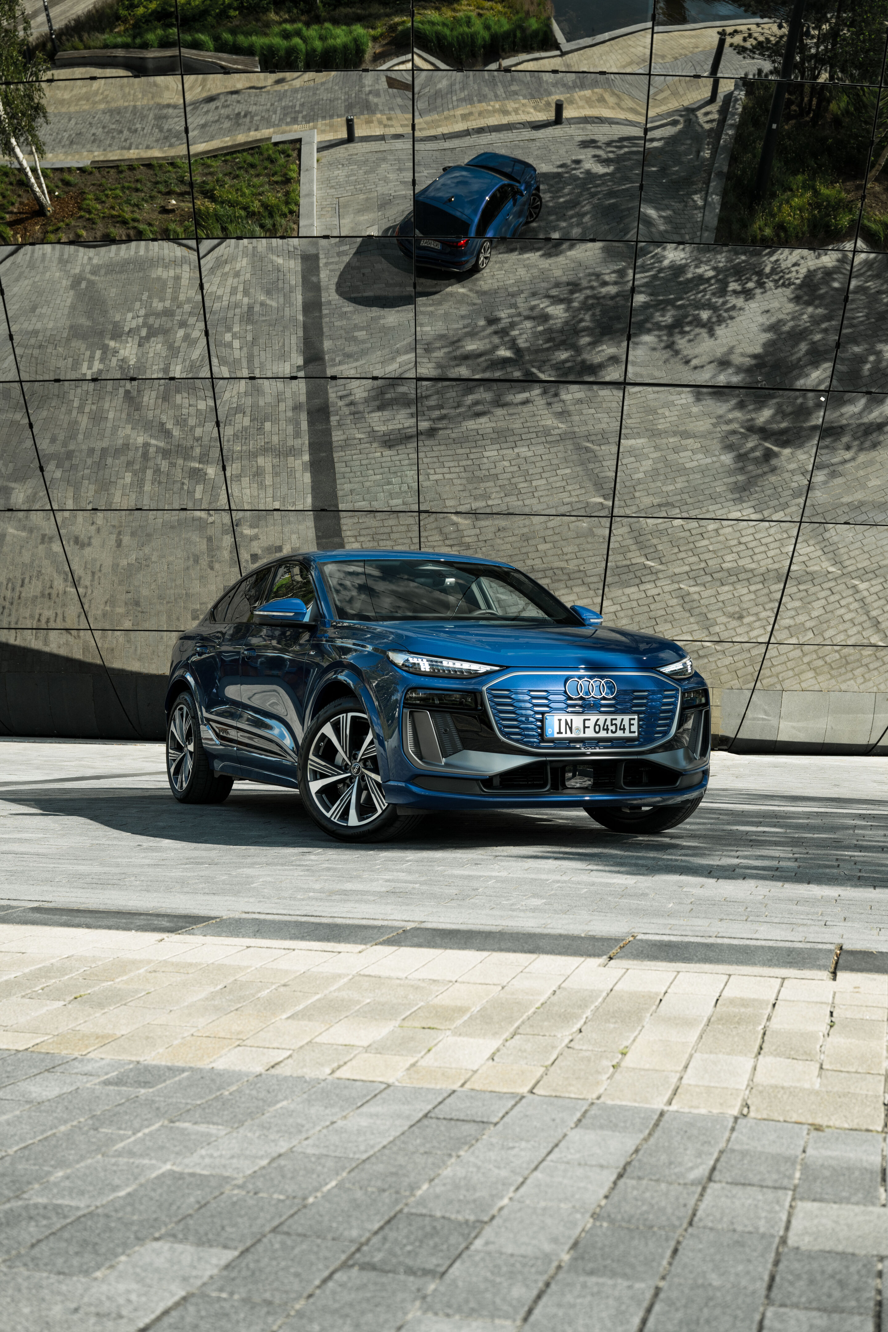 Audi Q6 e-tron Lifecycle Experience - Standaufnahme: Audi Q6 e-tron (Ascariblau), schräg von vorn aufgenommen, vor einer spiegelnden Glasfassade.