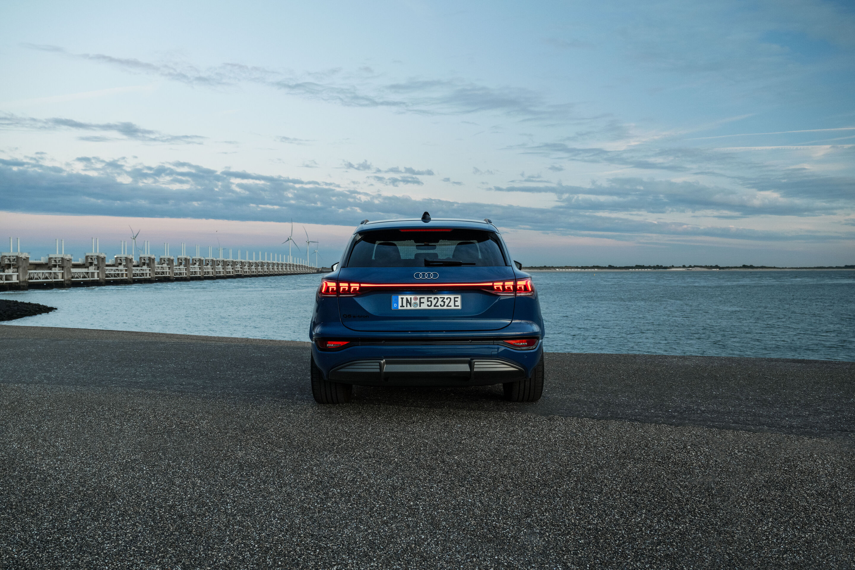 Audi Q6 e-tron Lifecycle Experience - Standaufnahme: Audi Q6 e-tron (Ascariblau), Heckansicht mit durchgehendem LED-Leuchtenband, geparkt an einer Küstenpromenade.