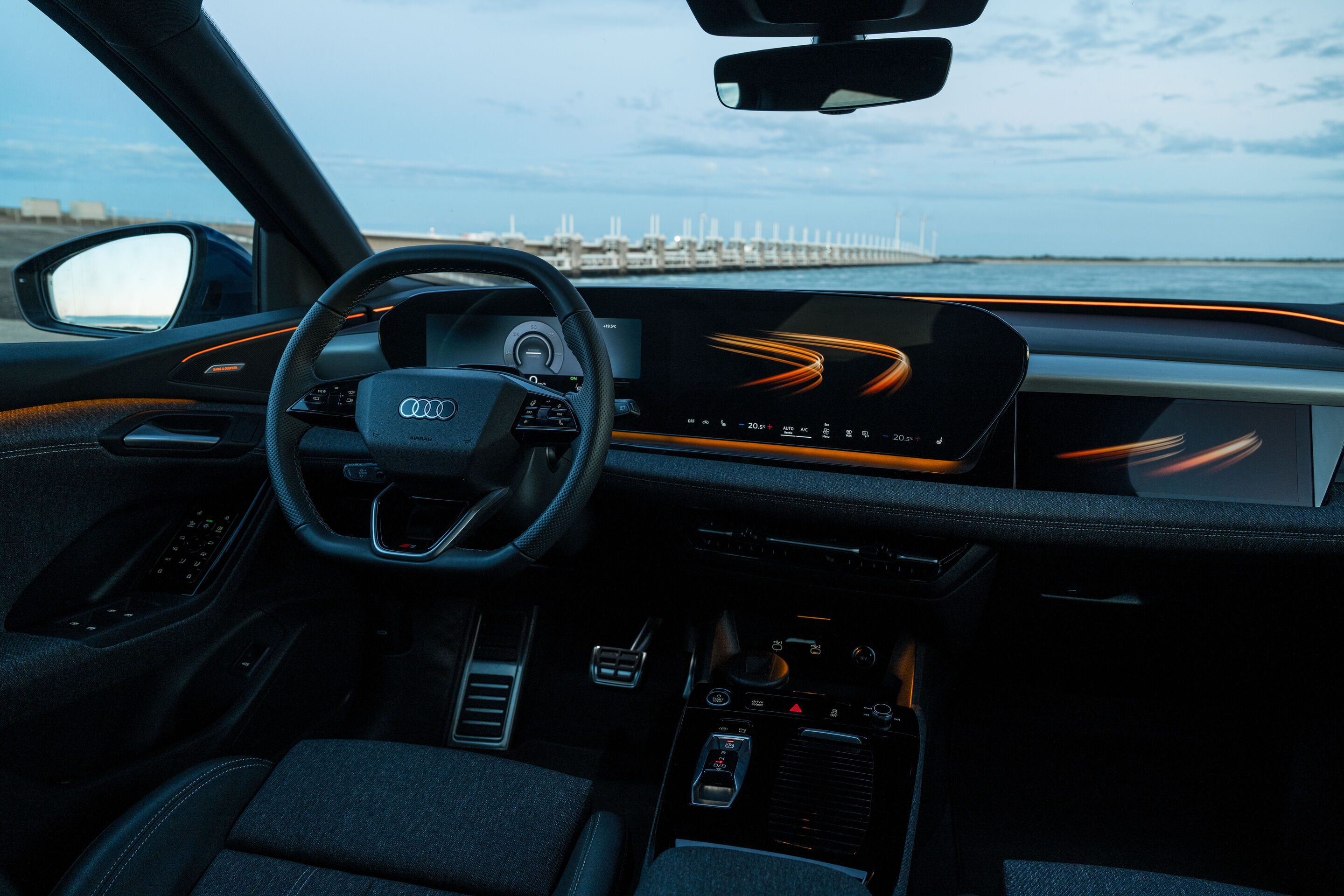 Audi Q6 e-tron Lifecycle Experience - Interior of the Audi Q6 e-tron showing one of the „experience worlds”, the steering wheel, digital cockpit and continuous display, highlighted by orange ambient lighting.