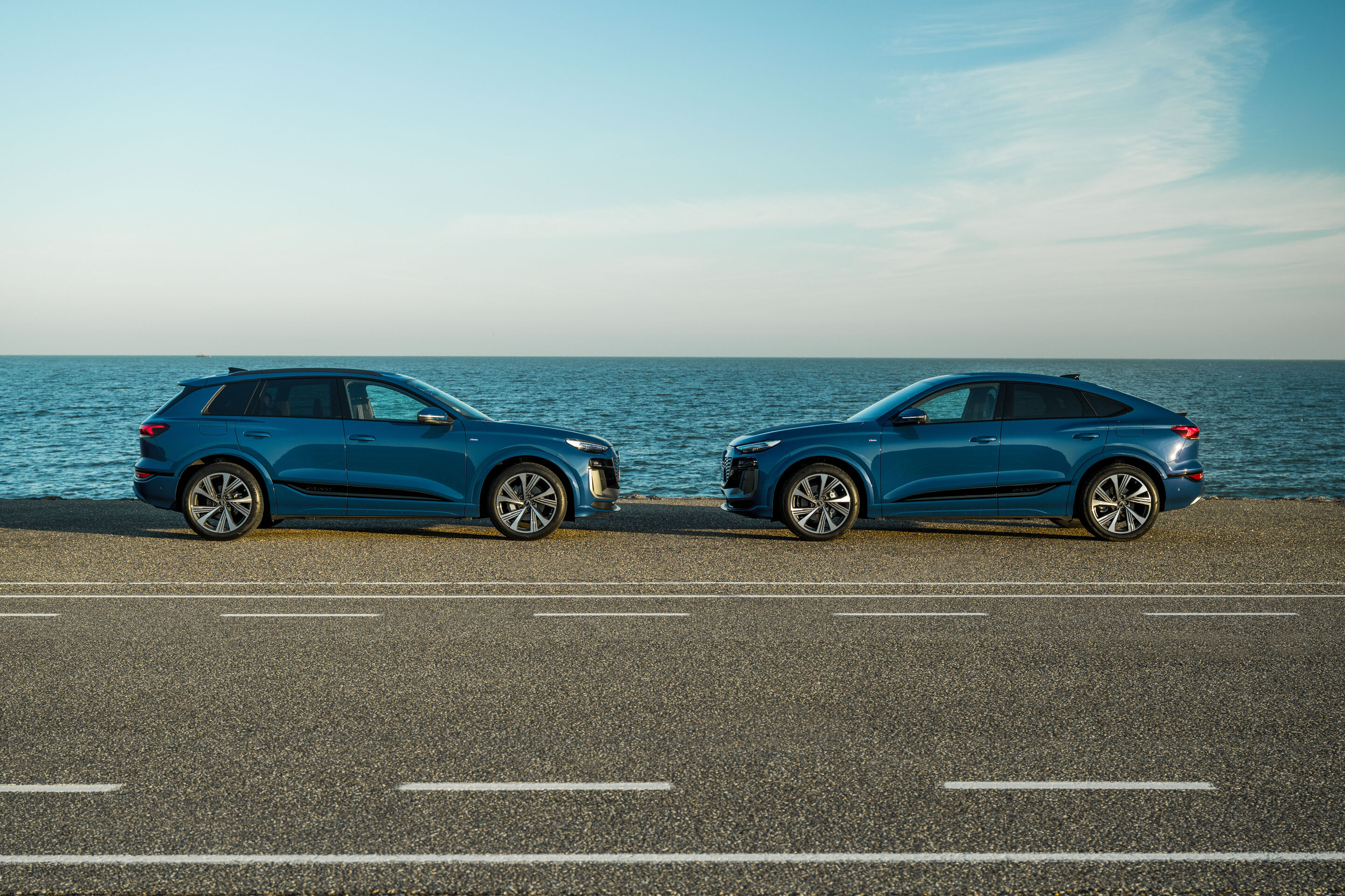 Audi Q6 e-tron Lifecycle Experience - Standaufnahme: Zwei Audi Q6 e-tron (Ascariblau), SUV und Sportback, in seitlicher Profilansicht gegenüberstehend auf einer Straße entlang der Küste mit offenem Meer im Hintergrund.