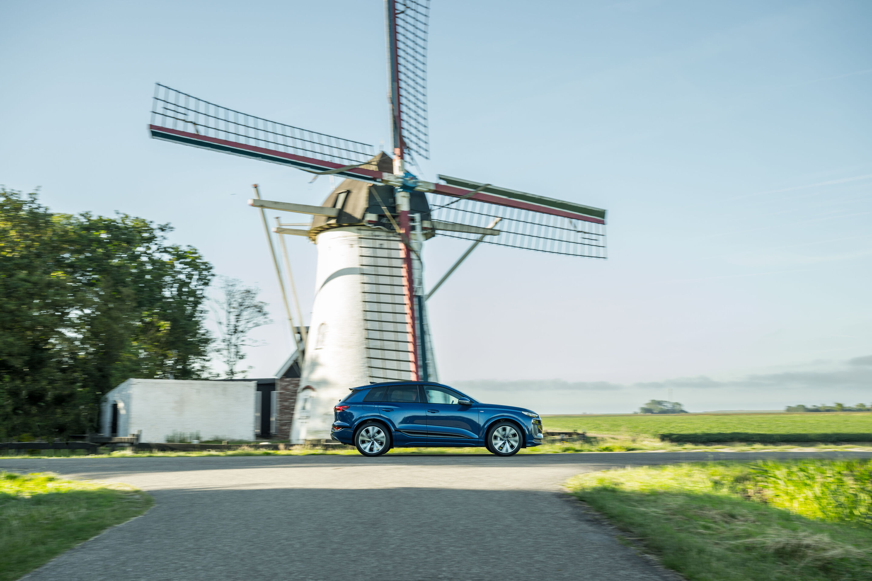 Audi Q6 e-tron Lifecycle Experience - Dynamische Aufnahme: Audi Q6 e-tron (Ascariblau), Seitenansicht in Bewegung auf einer Landstraße, vorbeifahrend an einer traditionellen Windmühle