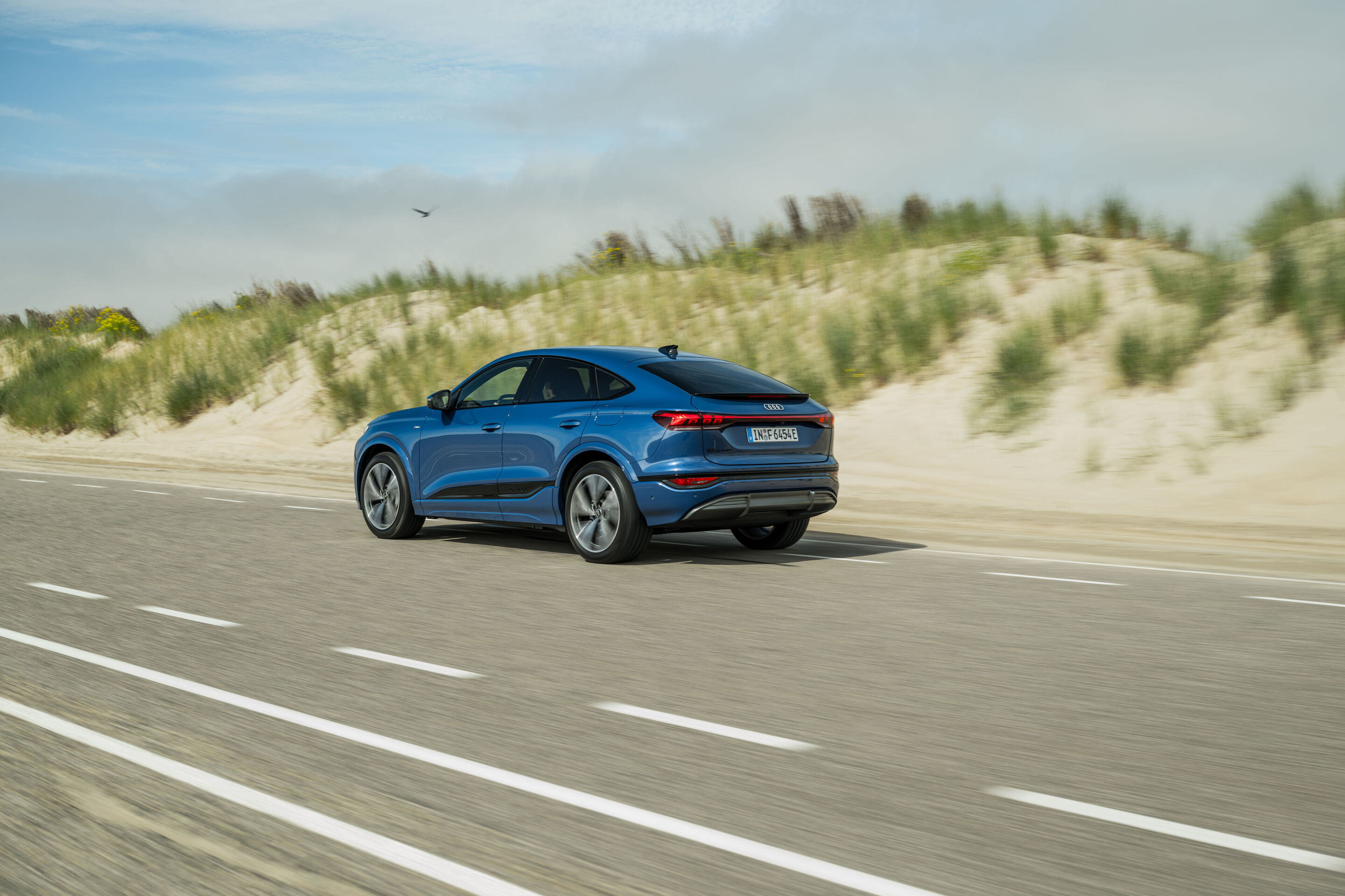 Audi Q6 e-tron Lifecycle Experience - Dynamische Aufnahme: Audi Q6 e-tron Sportback (Ascariblau), Heck-Dreiviertelansicht in Bewegung auf einer Küstenstraße, mit sandigen Dünen und Grasbewuchs im Hintergrund.