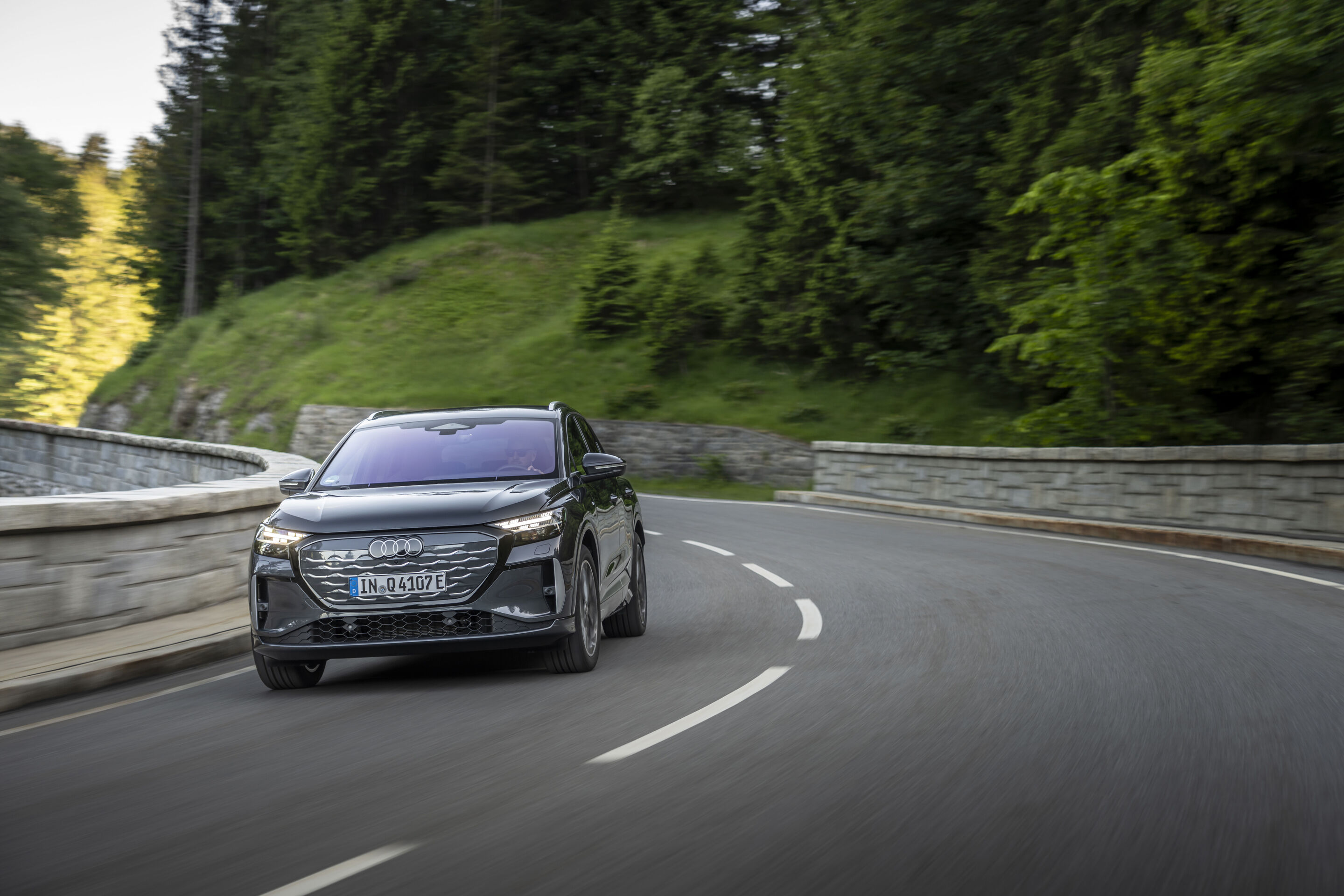 Ein Auto für jeden Moment: Unterwegs im Audi Q4 e-tron - Audi Q4 SUV e-tron in Tamboragrau, Outdoor-Fahraufnahme, Frontansicht auf kurvenreicher Straße in bewaldeter Landschaft.