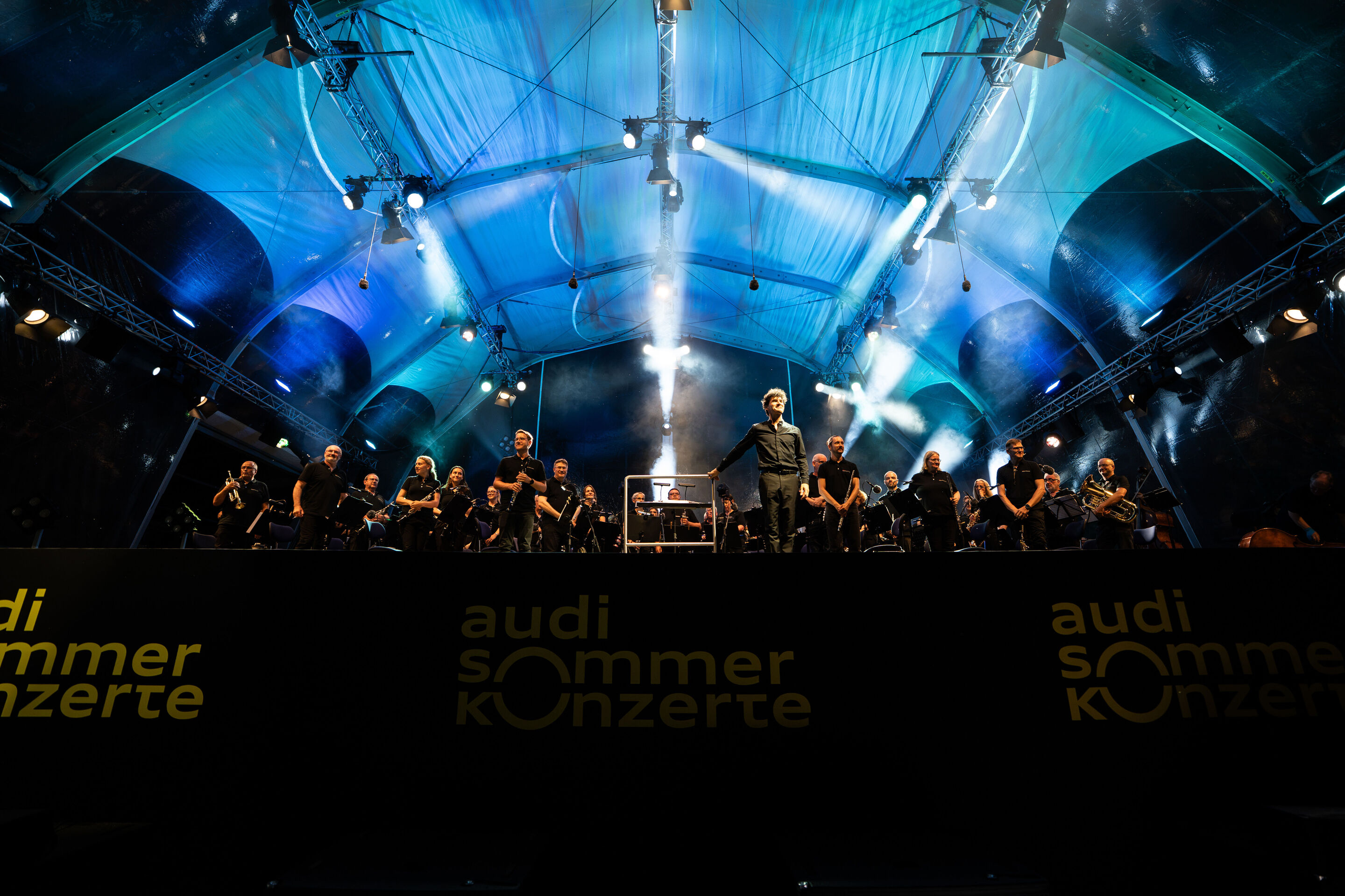 The Audi Wind Orchestra at the opening concert/classical open-air concert in Klenzepark - The Audi Wind Philharmonic on stage at the 2026 Classical Open-Air Concert in Klenzepark with conductor Mathias Wehr at the center of the image in front of the orchestra.