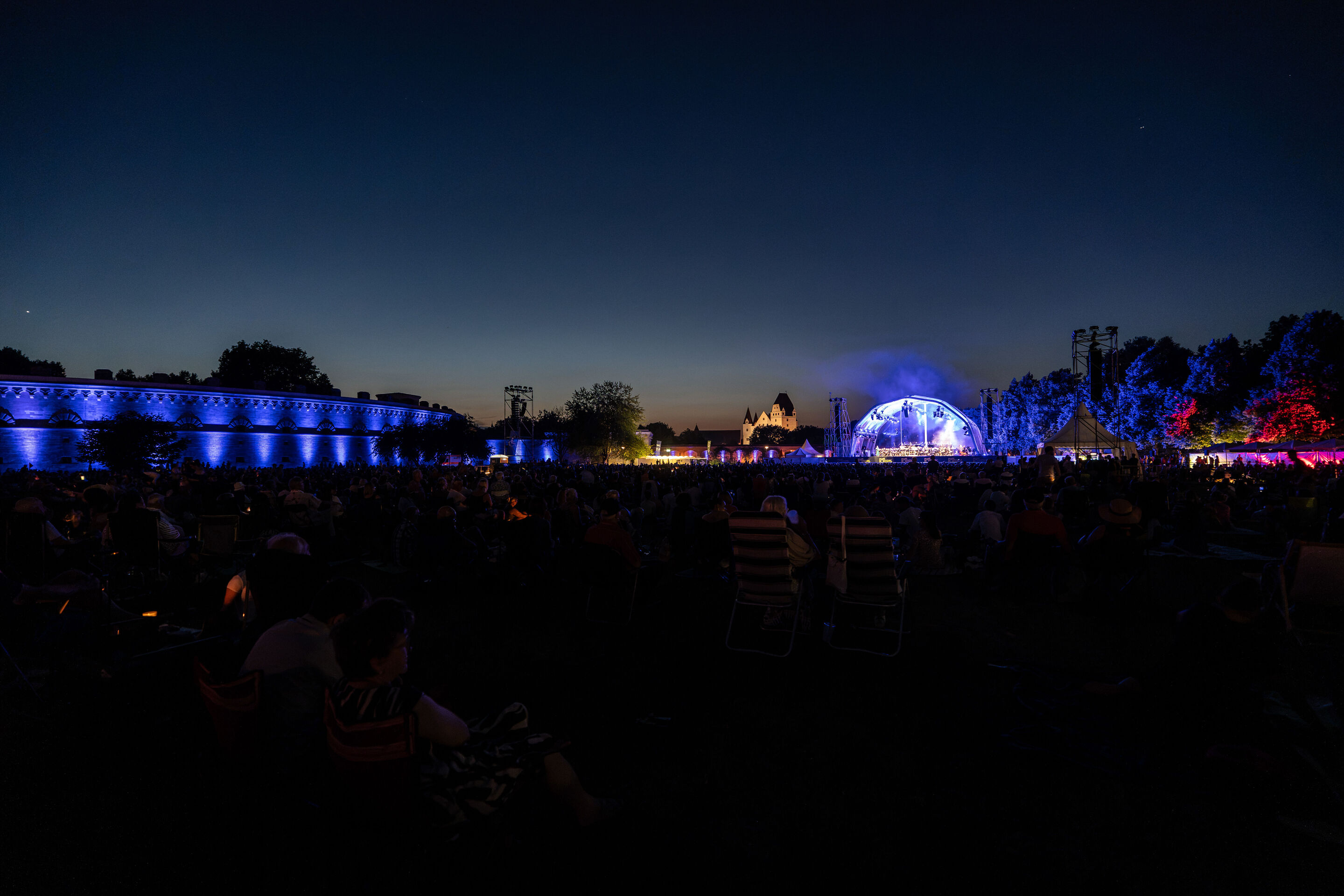 The Audi Wind Orchestra at the opening concert/classical open-air concert in Klenzepark - View across the Klenzepark meadow at night towards the illuminated open-air stage; on the left, the Reduit Tilly is lit in blue, with the skyline of Ingolstadt’s...