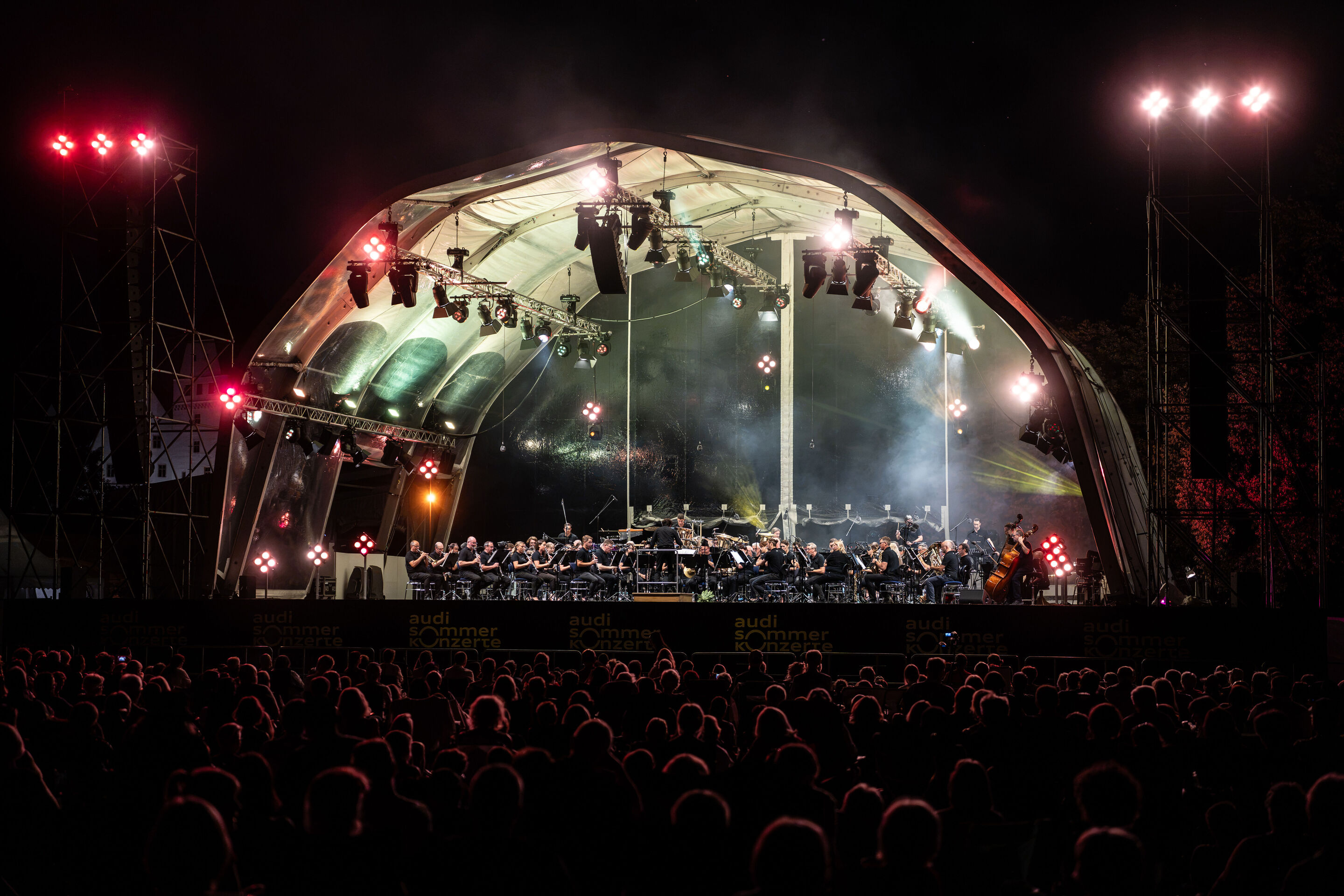 The Audi Wind Orchestra at the opening concert/classical open-air concert in Klenzepark - View from the audience towards the illuminated open-air stage in Klenzepark under a dark night sky: The Audi Wind Philharmonic performs beneath the large-scale stage...
