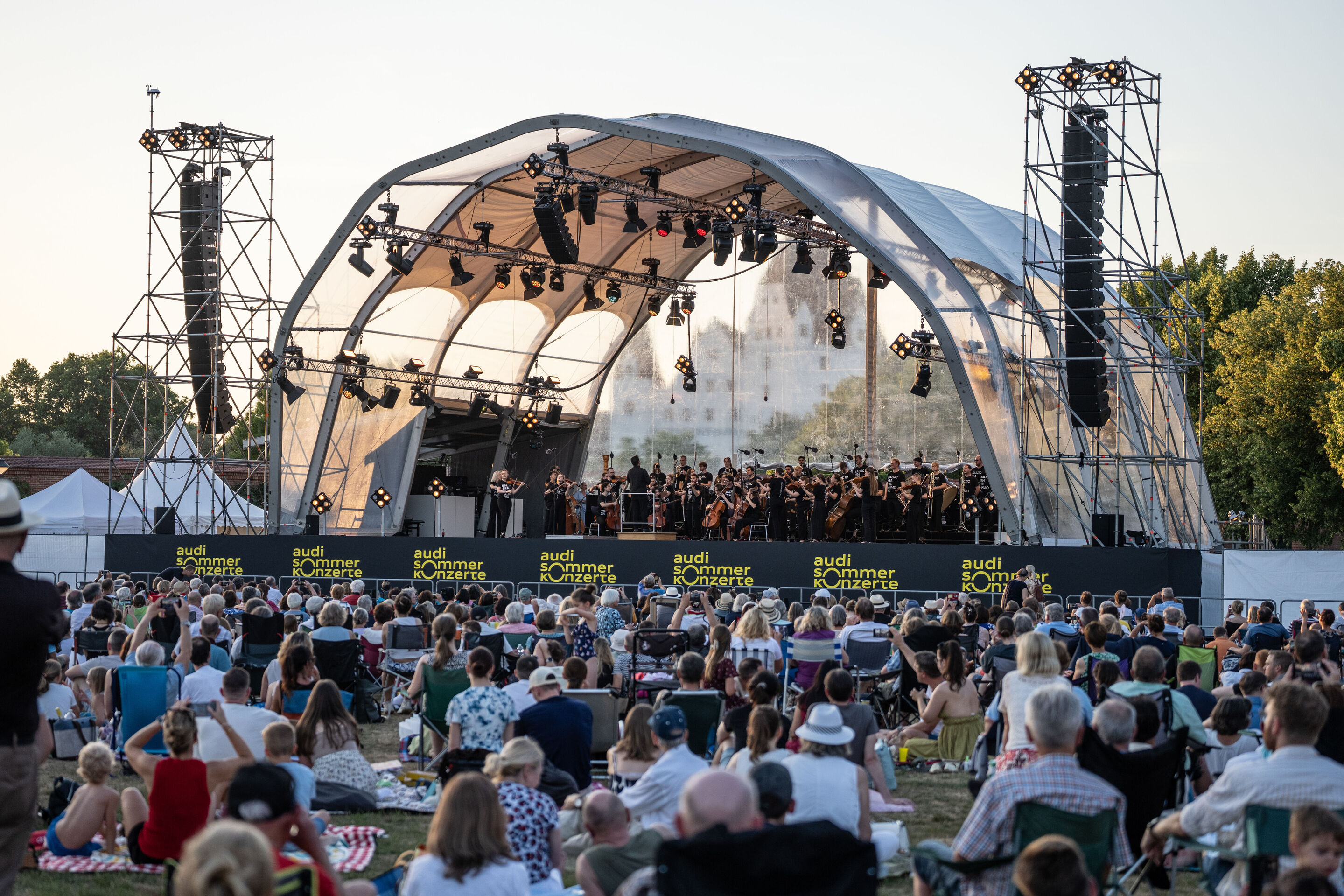 Baltic Sea Philharmonic performing at the classical open-air concert in Klenzepark - View from the audience towards the stage of the 2026 Classical Open-Air Concert in Klenzepark: Baltic Sea Philharmonic performs beneath the open-air stage structure,...