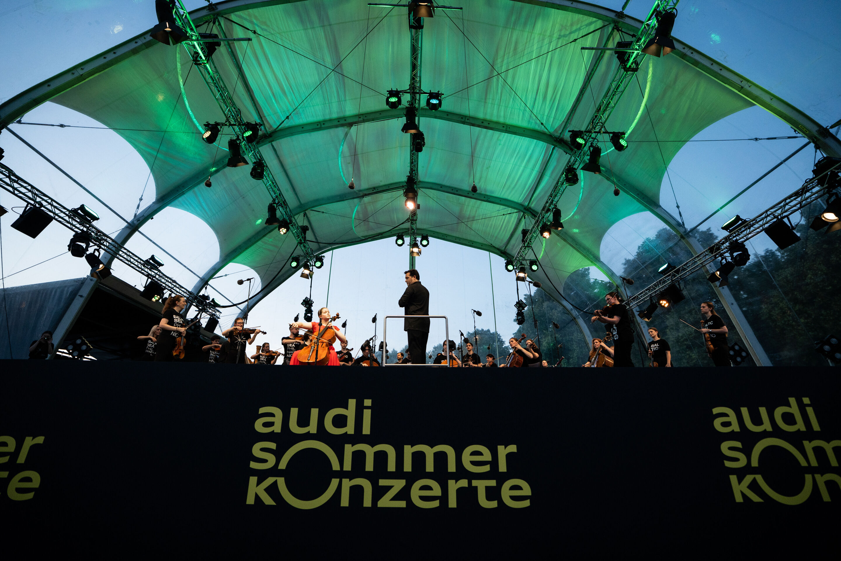 Baltic Sea Philharmonic performing at the classical open-air concert in Klenzepark - View from the audience towards the stage of the 2026 Classical Open-Air Concert in Klenzepark: Under the green-lit stage structure, the musicians of the Baltic Sea Philharmonic...