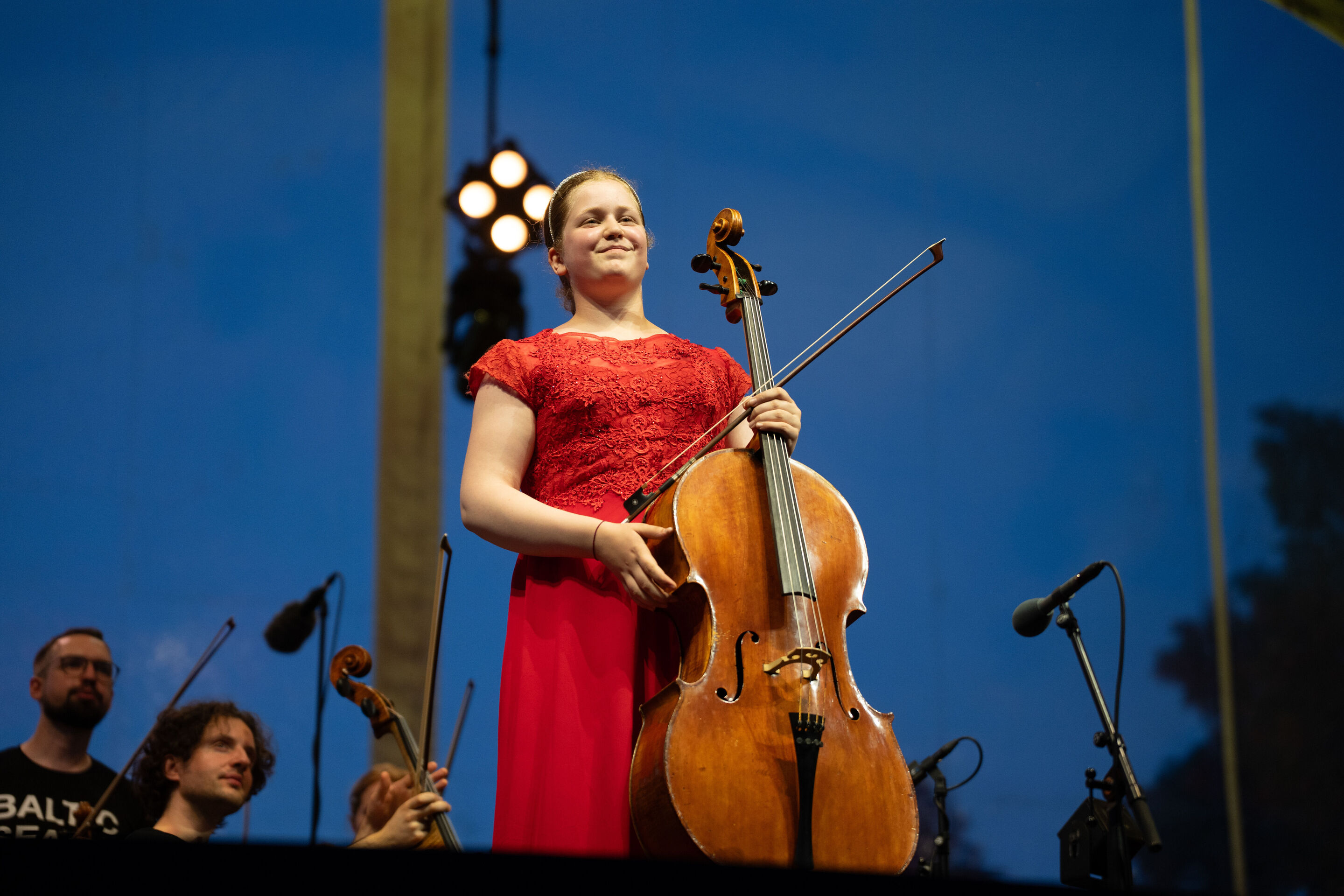 Baltic Sea Philharmonic performing at the classical open-air concert in Klenzepark - Cellist Charlotte Melkonian stands on stage with her cello at the 2026 Classical Open-Air Concert in Klenzepark, while members of the Baltic Sea Philharmonic are visible...