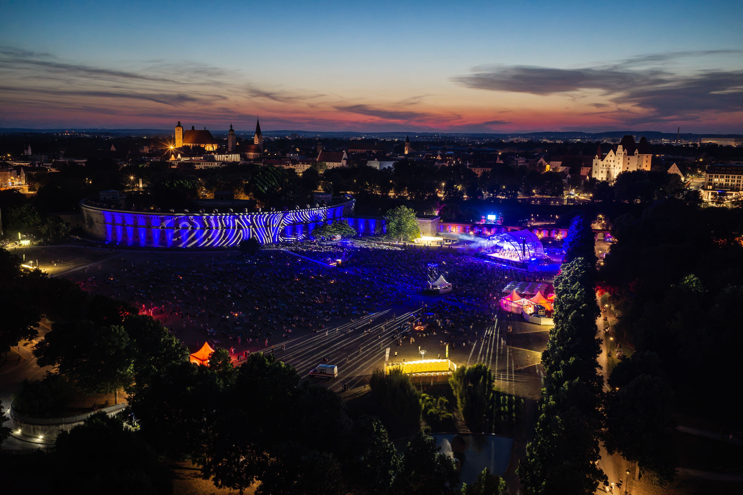 Baltic Sea Philharmonic performing at the classical open-air concert in Klenzepark - Aerial view of the 2026 Classical Open-Air Concert in Klenzepark, showing the blue-lit concert stage, the illuminated Reduit Tilly, and the historic old town of Ingolstadt...