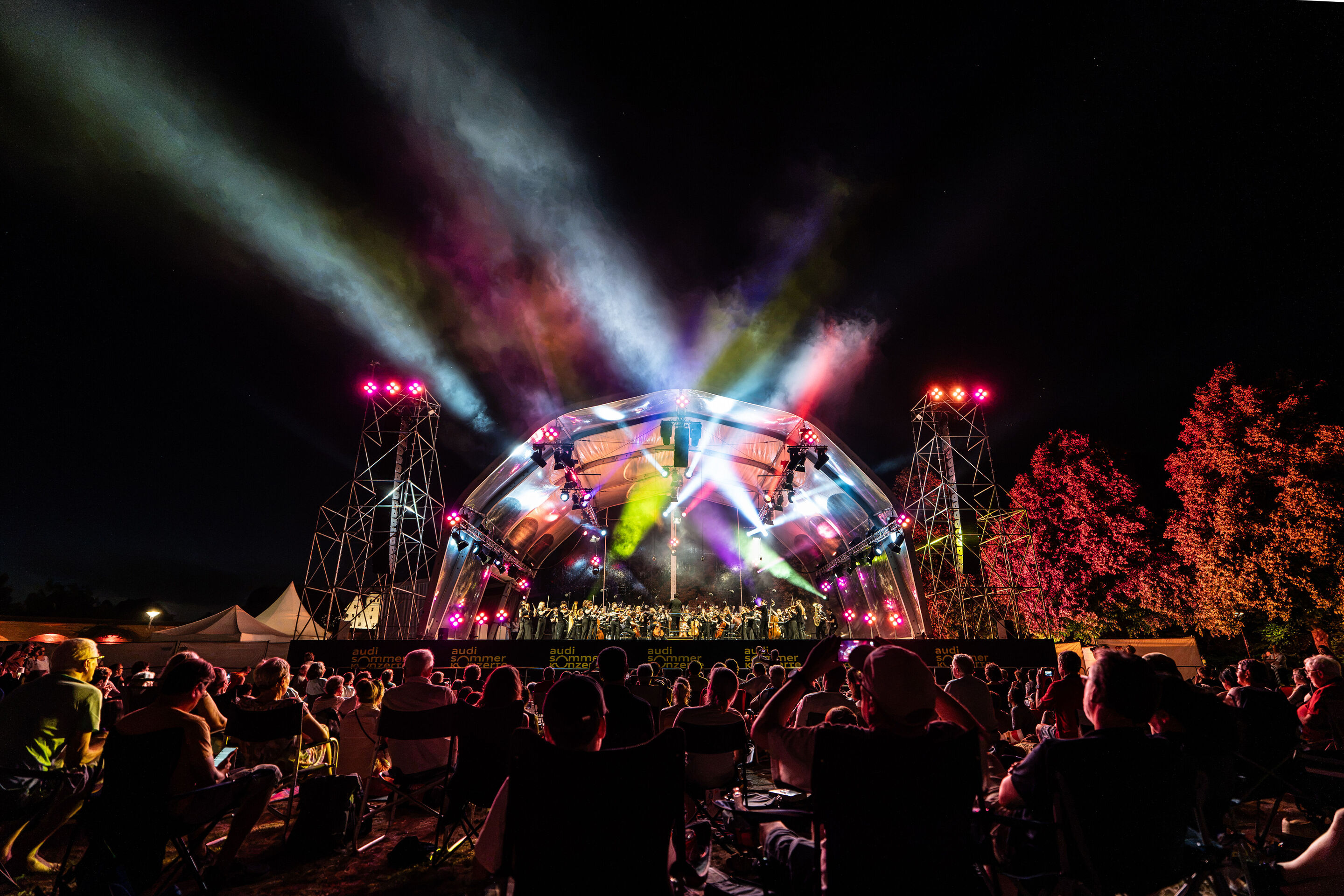 Baltic Sea Philharmonic performing at the classical open-air concert in Klenzepark - View from the audience towards the big concert stage of the 2026 Classical Open-Air Concert in Klenzepark, illuminated by coloured light installations, while visitors...